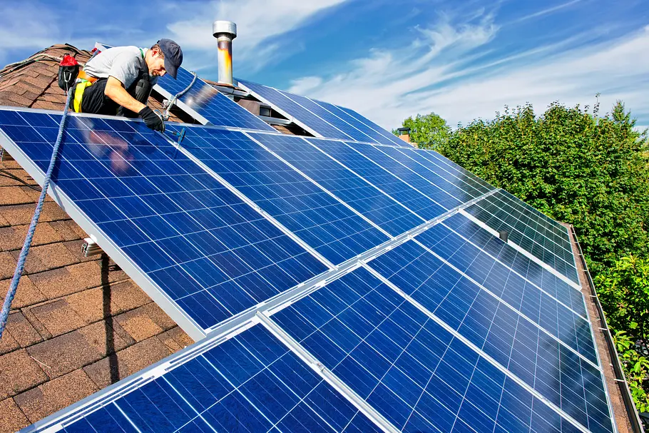 solaranlage-selbst-installieren-was-ist-erlaubt Eine Person bringt Solarpanels auf einem Dach an, die Panels glänzen bläulich, es sind grüne Bäume und Blauer Himmel zu sehen