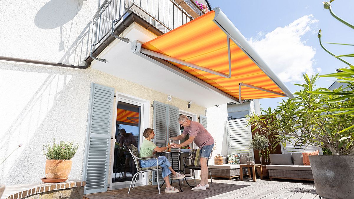 Eine ausgefahrene gelb-orange gestreifte Markise, die unter einem Balkon angebracht ist. Ein Mann und eine Frau sitzen darunter im Schatten.