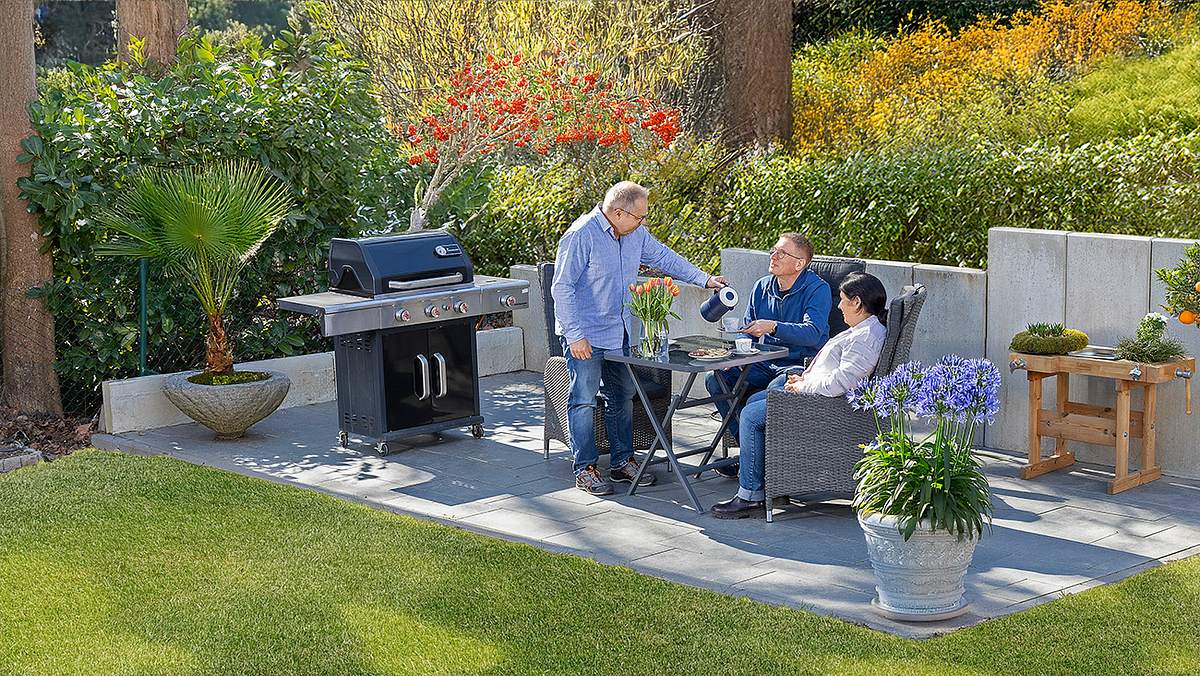 Auf einer grau gepflasterten Terrasse stehen ein Tisch mit drei Stühlen zentral, links davon ein Gasgrill, rechts ein Pflanztisch hinten und im Vordergrund ein großer Blumentopf aus Stein mit blauen Blumen. Auf den Stühlen sitzen eine Frau rechts und ein Mann mittig. Links steht ein Mann, der dem sitzenden Mann Kaffee aus einer Kanne einschenkt. Auf dem Tisch befinden sich noch Plätzchen und Blumen in einer Glasvase. Im Hintergrund sieht man eine halbhohe Mauer aus Stahlbeton, dahinter ein Hang mit vielen Blumen und Sträuchern.