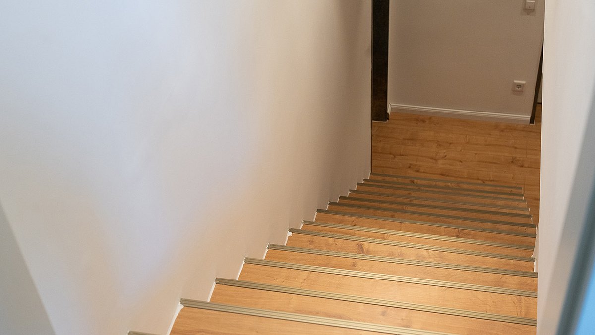 Eine gerade verlaufende Treppe mit Holzstufen und Metallkanten führt zu einem Flur mit hellem Holzboden.