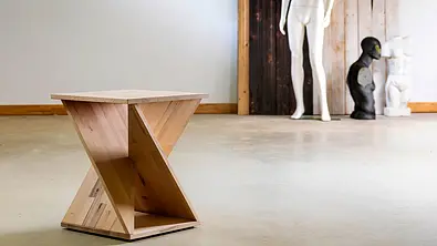 Ein Hocker aus hellem Holz in verdrehter Form steht auf einem Betonboden, im Hintergrund befinden sich drei Torso-Skulpturen. - Foto: sidm / Michael Müller-Münker Ein Hocker aus hellem Holz in verdrehter Form steht auf einem Betonboden, im Hintergrund befinden sich drei Torso-Skulpturen. - Foto: sidm / Michael Müller-Münker