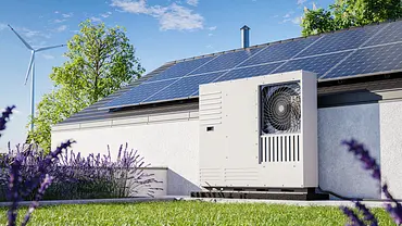 Rasen vor einem Haus, eine große Wärmepumpe ist erkennbar, auf dem Dach sind Photovoltaik-Paneele. Der Himmel ist blau, im Hintergrund ist ein Windrad. - Foto: istockphoto.com / guteksk7 Rasen vor einem Haus, eine große Wärmepumpe ist erkennbar, auf dem Dach sind Photovoltaik-Paneele. Der Himmel ist blau, im Hintergrund ist ein Windrad. - Foto: istockphoto.com / guteksk7
