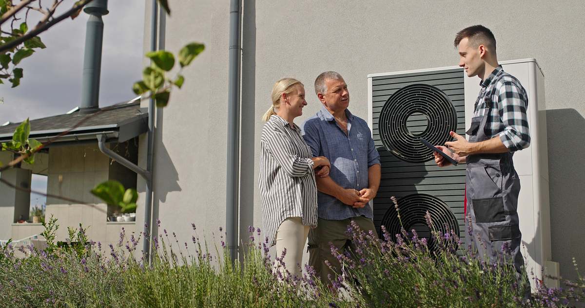 Drei Personen stehen neben einer Wärmepumpe an einer Hauswand. Ein Techniker in Arbeitskleidung erklärt einem älteren Ehepaar in Freizeitkleidung, wie ihre neue Heizung funktioniert. 