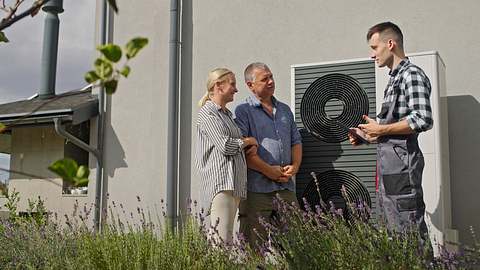 Drei Personen stehen neben einer Wärmepumpe an einer Hauswand. Ein Techniker in Arbeitskleidung erklärt einem älteren Ehepaar in Freizeitkleidung, wie ihre neue Heizung funktioniert.  - Foto: istockphoto.com / StockSeller_ukr