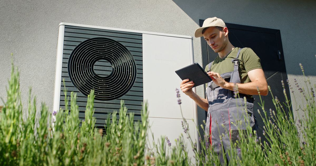 Techniker mit Tablet steht vor einer installierten Luft-Wärmepumpe an einem Wohngebäude, im Vordergrund blühender Lavendel.