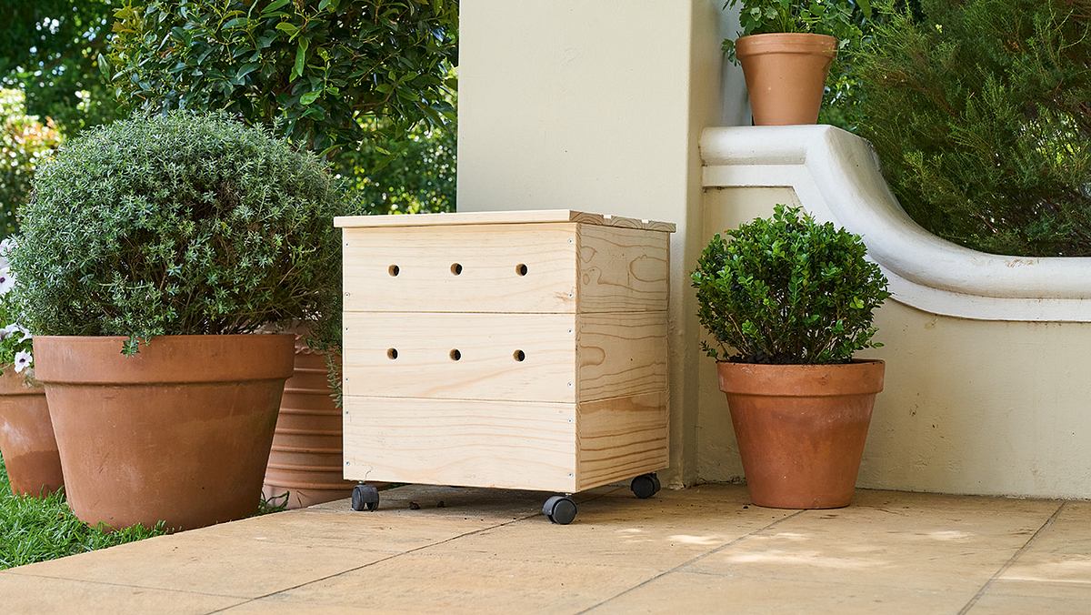 Eine Holzkiste mit Lüftungslöchern steht auf Rollen zwischen zwei Pflanzen in Tontöpfen auf einer überdachten Terrasse.